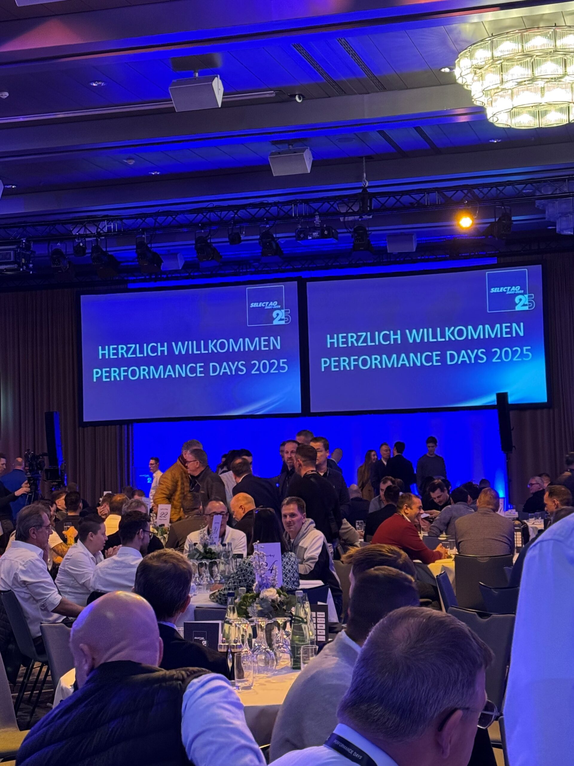 Veranstaltung „Performance Days 2025“ mit zahlreichen Gästen an runden Tischen in einem Konferenzsaal. Auf zwei Projektionswänden steht: „Herzlich Willkommen Performance Days 2025“.