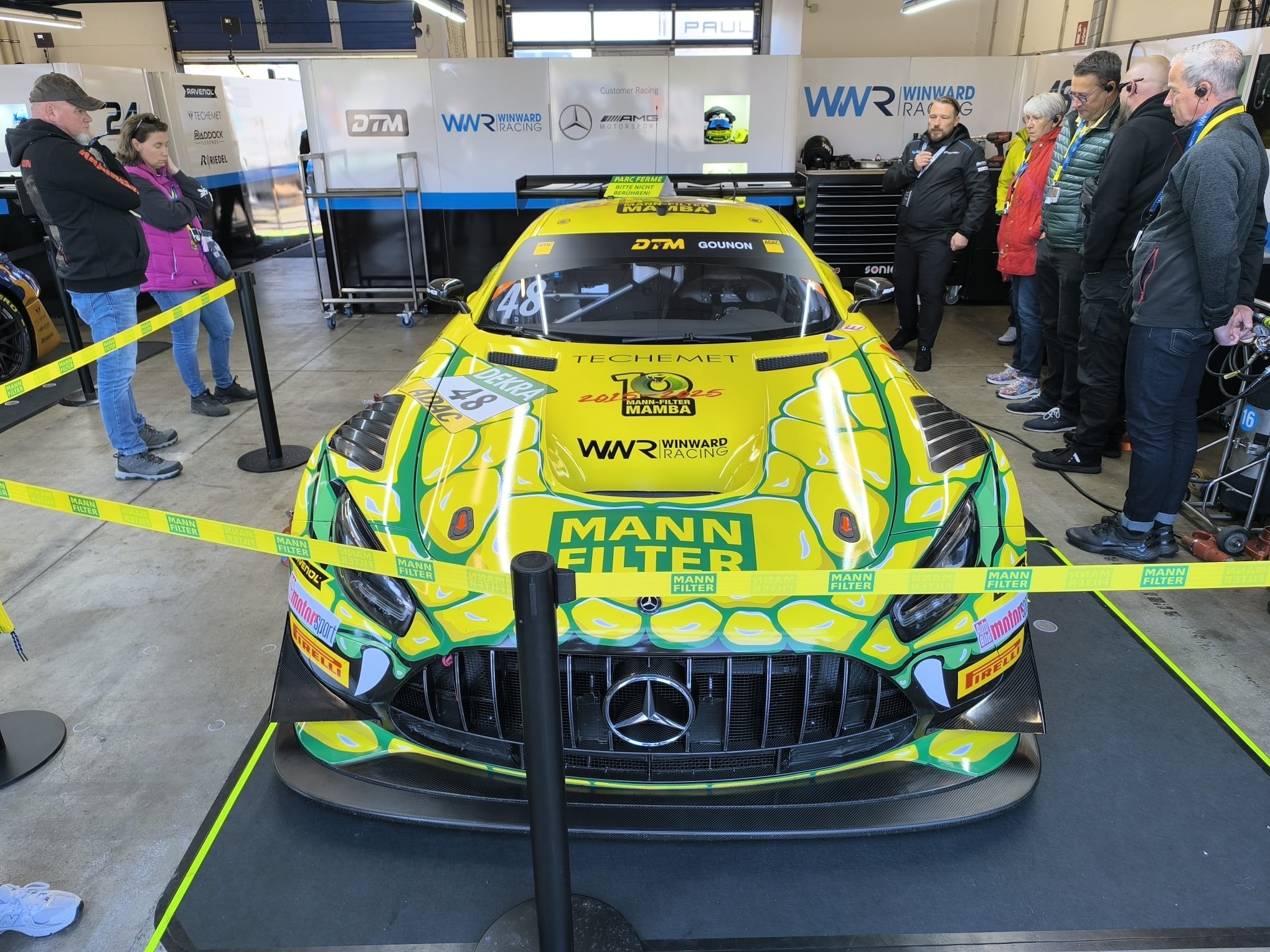 Gelber Mercedes-AMG GT3 von MANN FILTER im Fahrerlager, umgeben von Zuschauern. DTM-Branding und Logos von Sponsoren auf dem Fahrzeug.